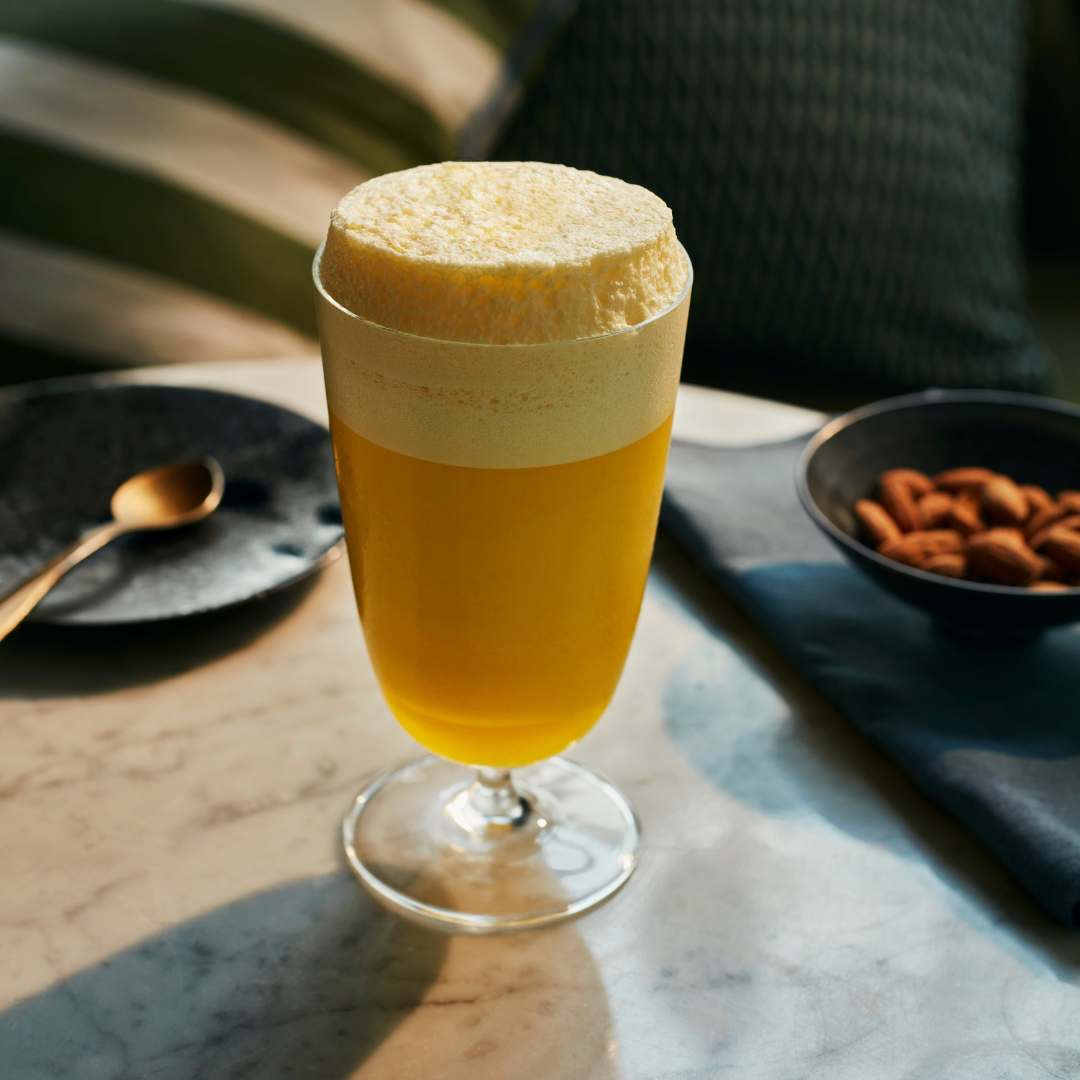 Glass of yellow drink with foam on a table with a spoon and bowl of nuts.
