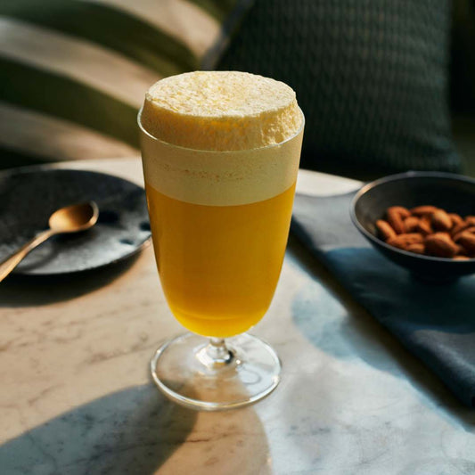 Glass of yellow drink with foam on a table with a spoon and bowl of nuts.