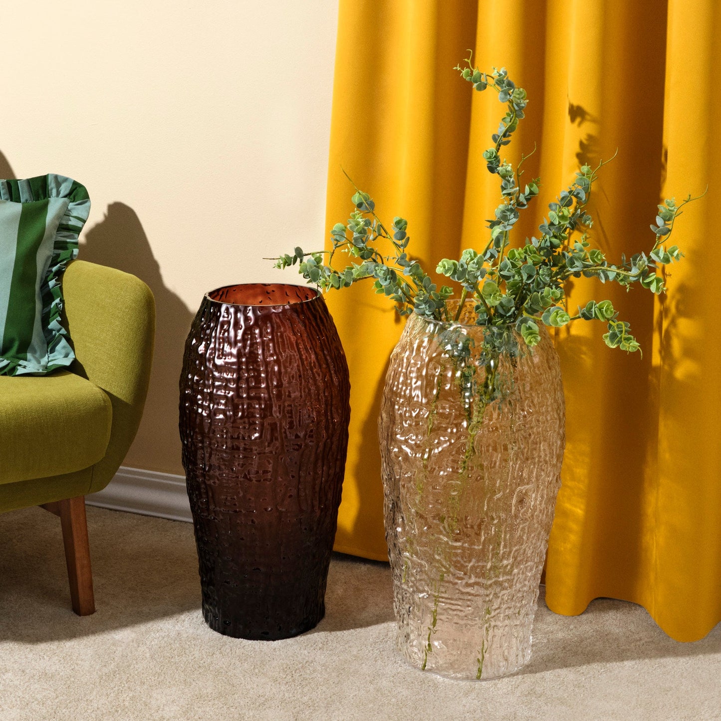 Two textured vases with greenery against a yellow curtain and green chair.