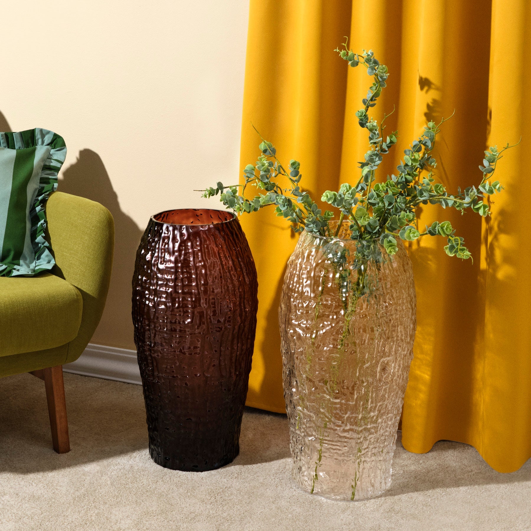 Two textured vases with greenery against a yellow curtain and green chair.