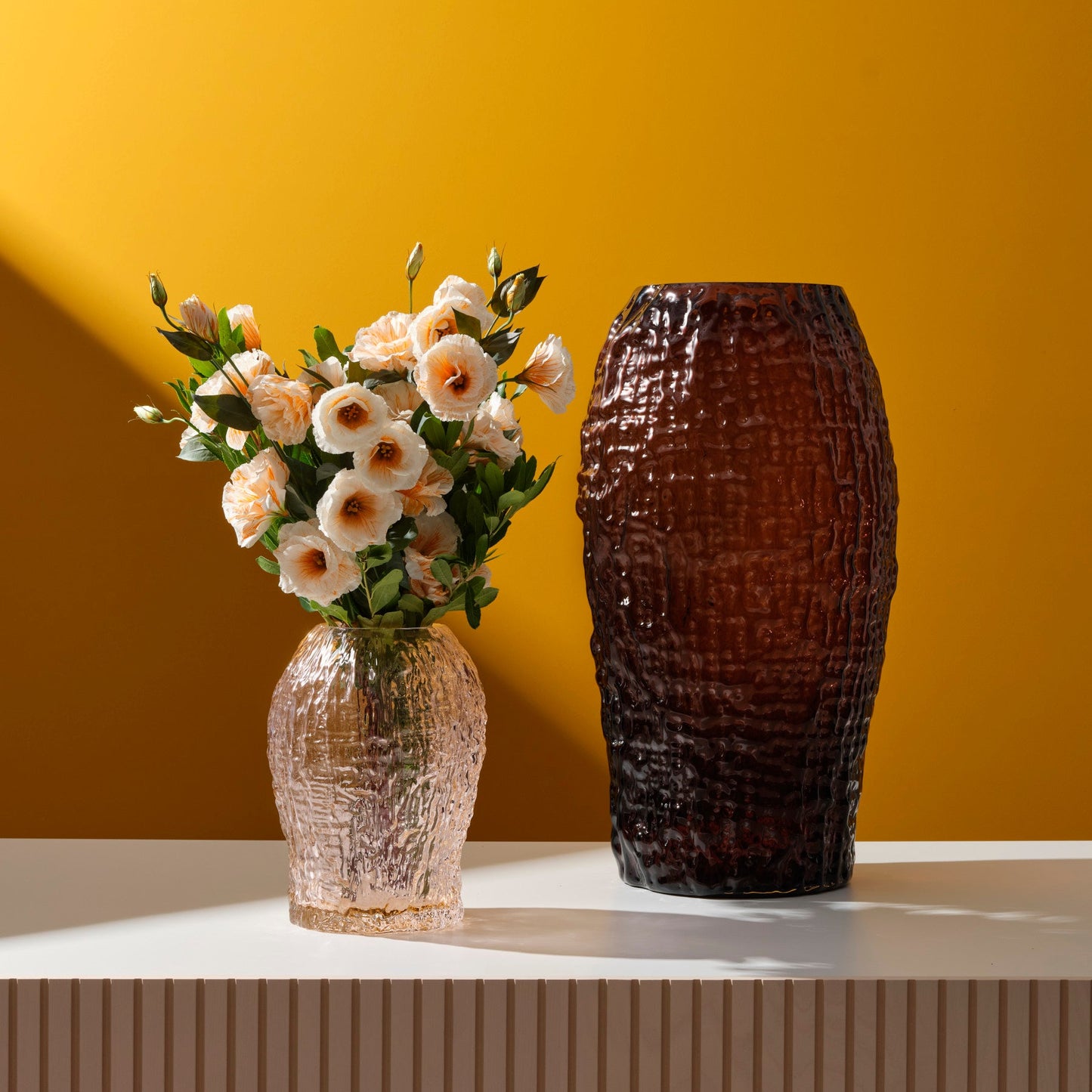 Two vases, one with flowers and the other textured, against a yellow wall.