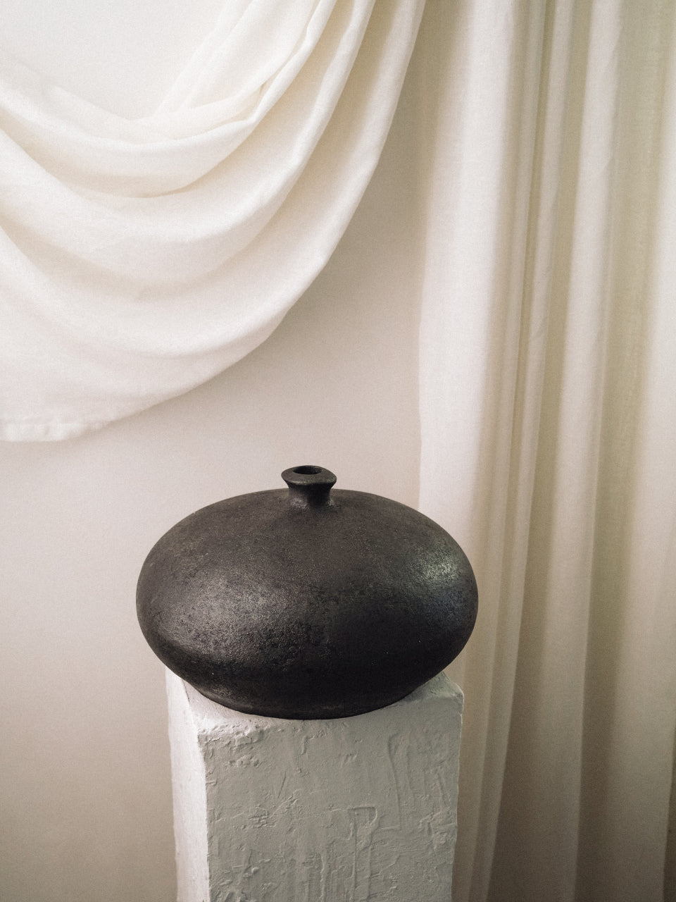 Black ceramic pot on a white pedestal with white draped fabric in the background