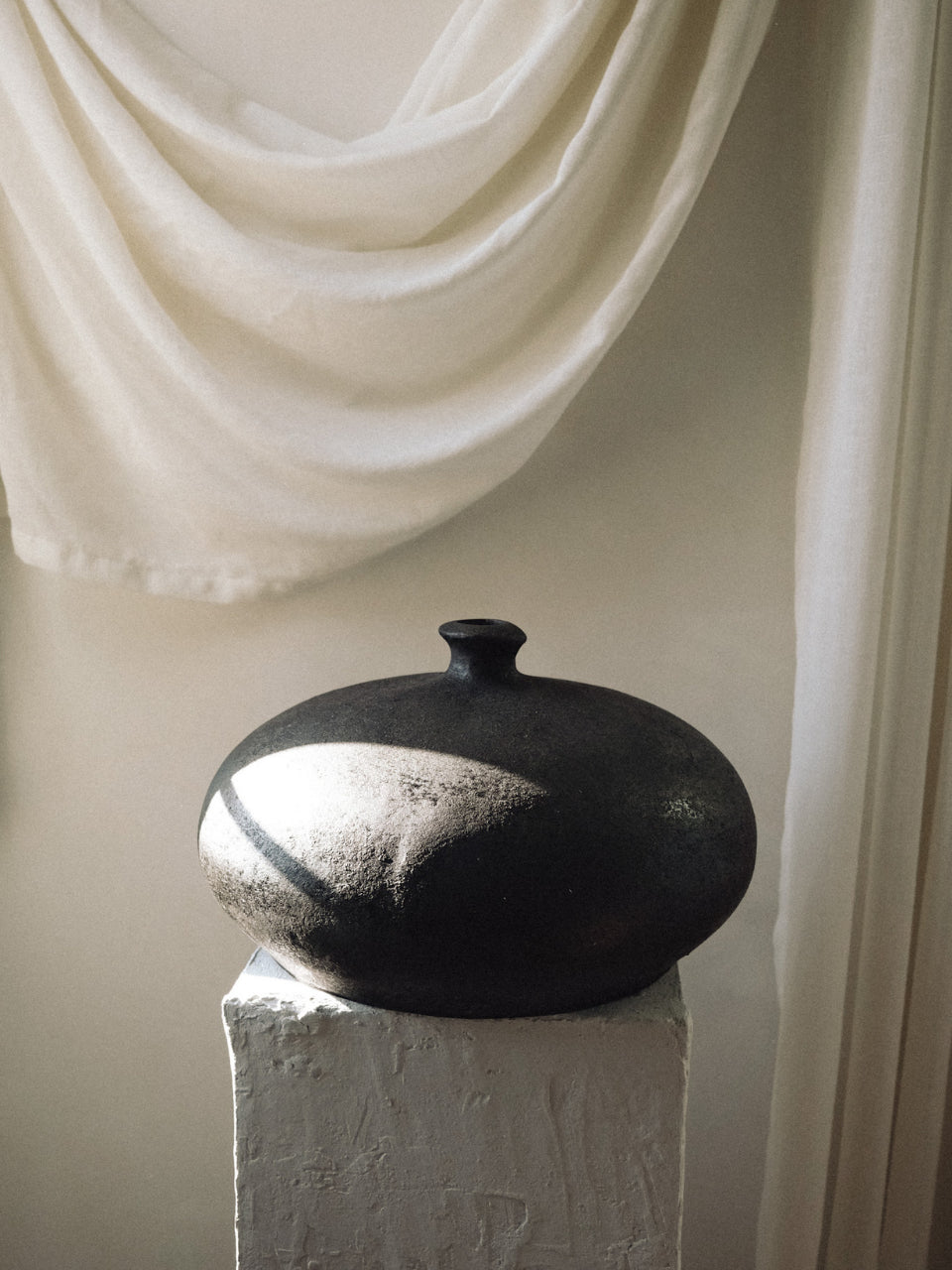 Black ceramic vase on a white pedestal with draped white fabric in the background