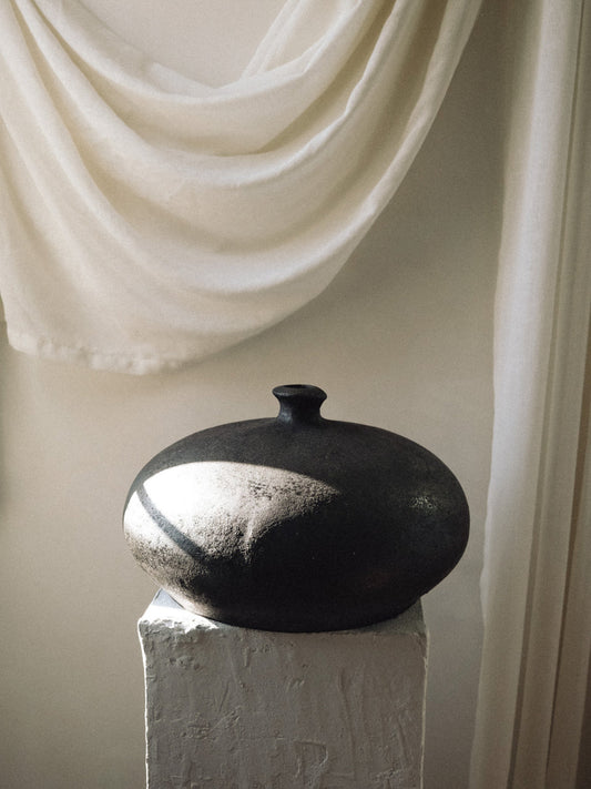 Black ceramic vase on a white pedestal with draped white fabric in the background