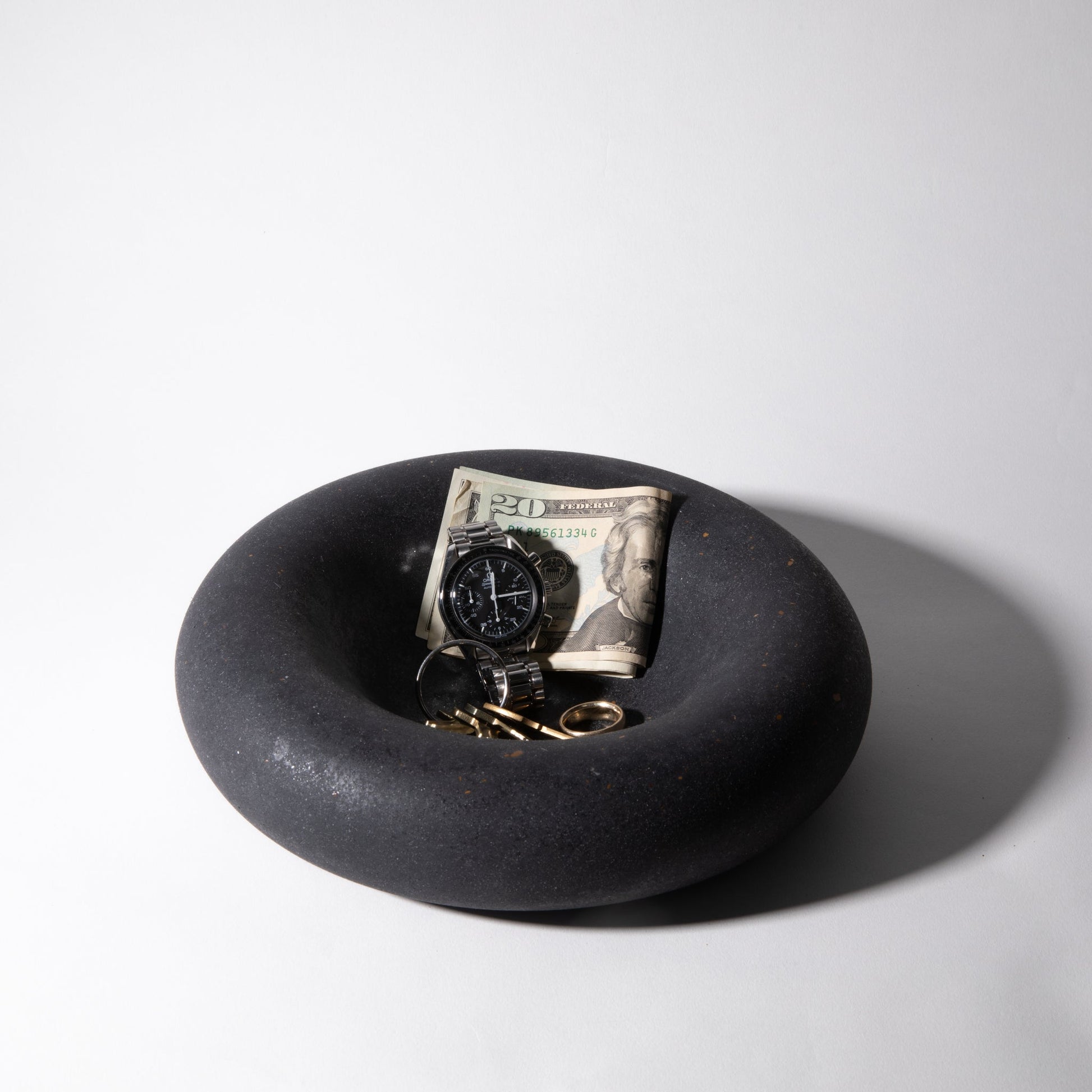 Black dish with a watch, keys, and dollar bill on a white background