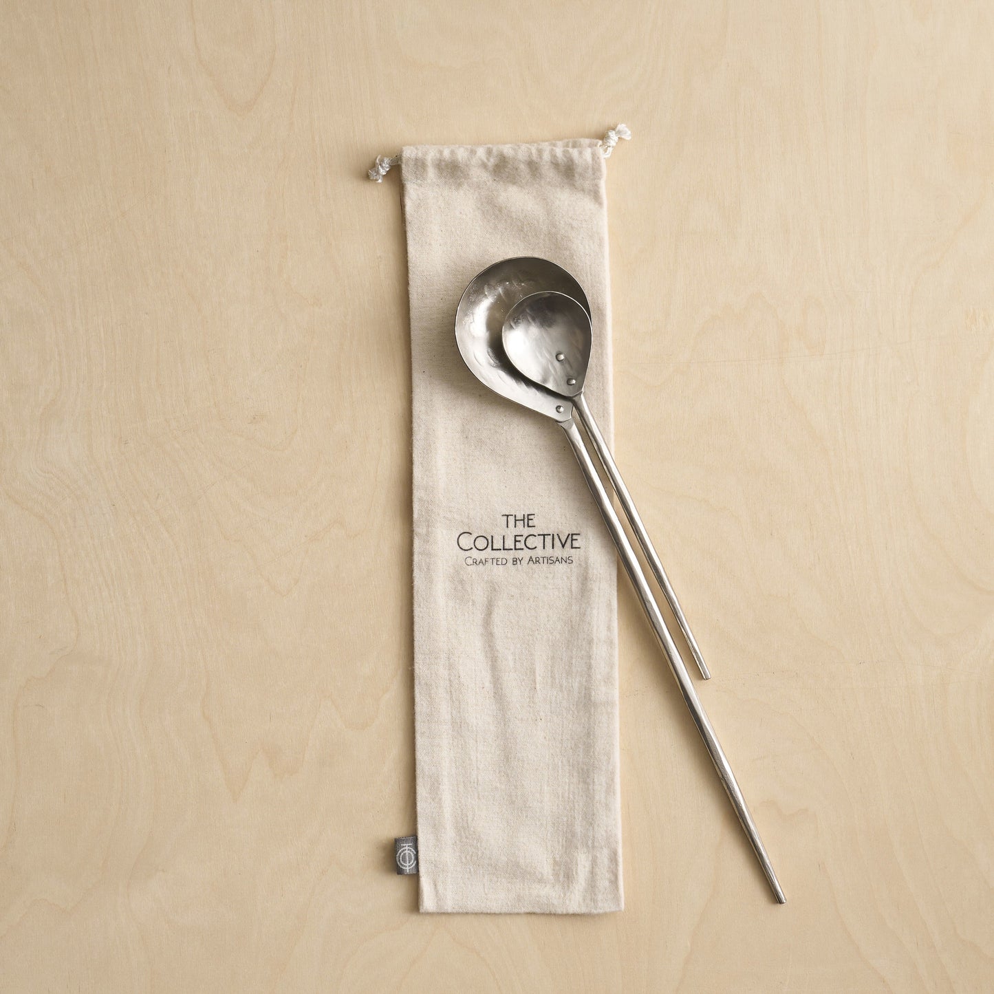 Metallic ladle and spoon on a beige fabric pouch with 'The Collective' branding against a beige background