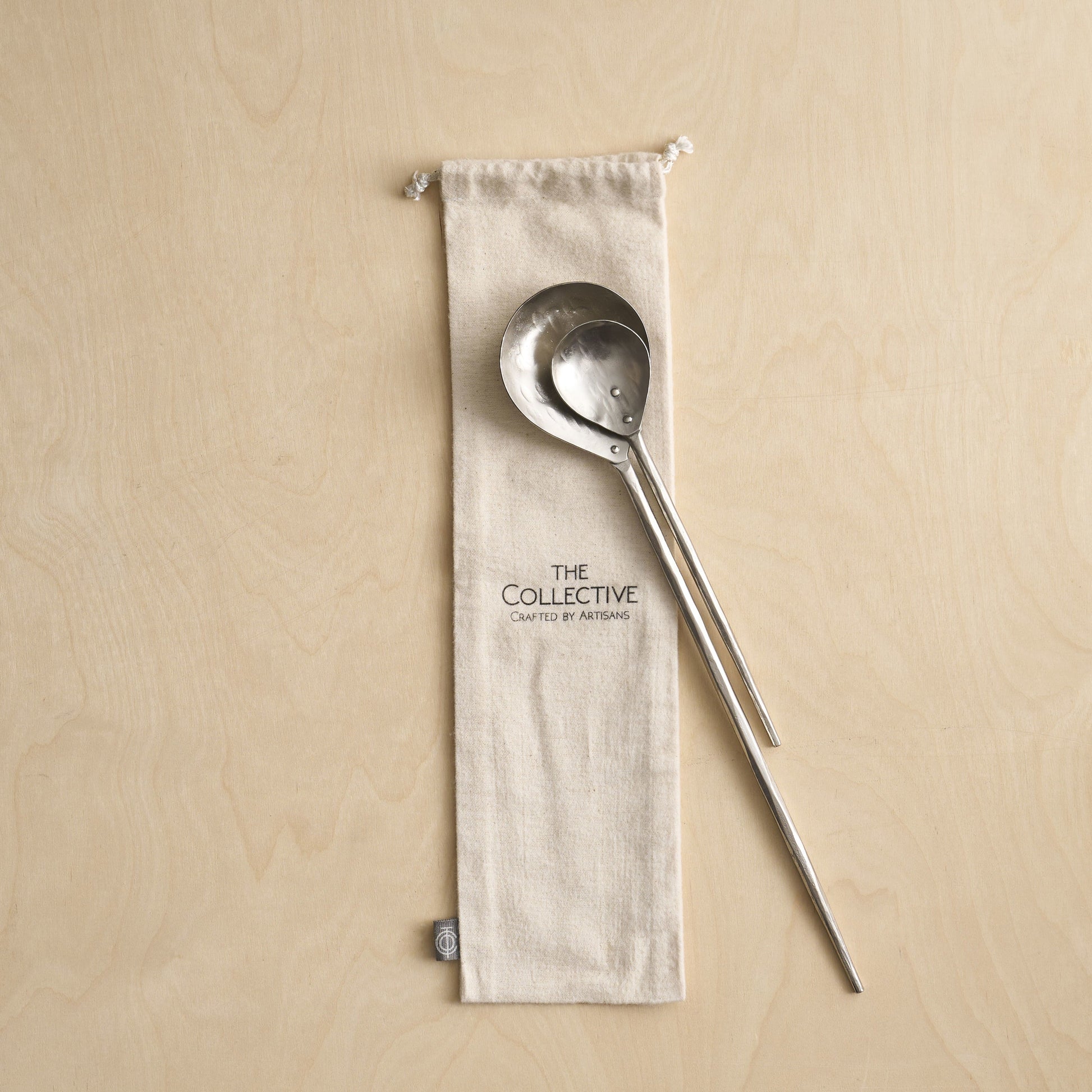 Metallic ladle and spoon on a beige fabric pouch with 'The Collective' branding against a beige background