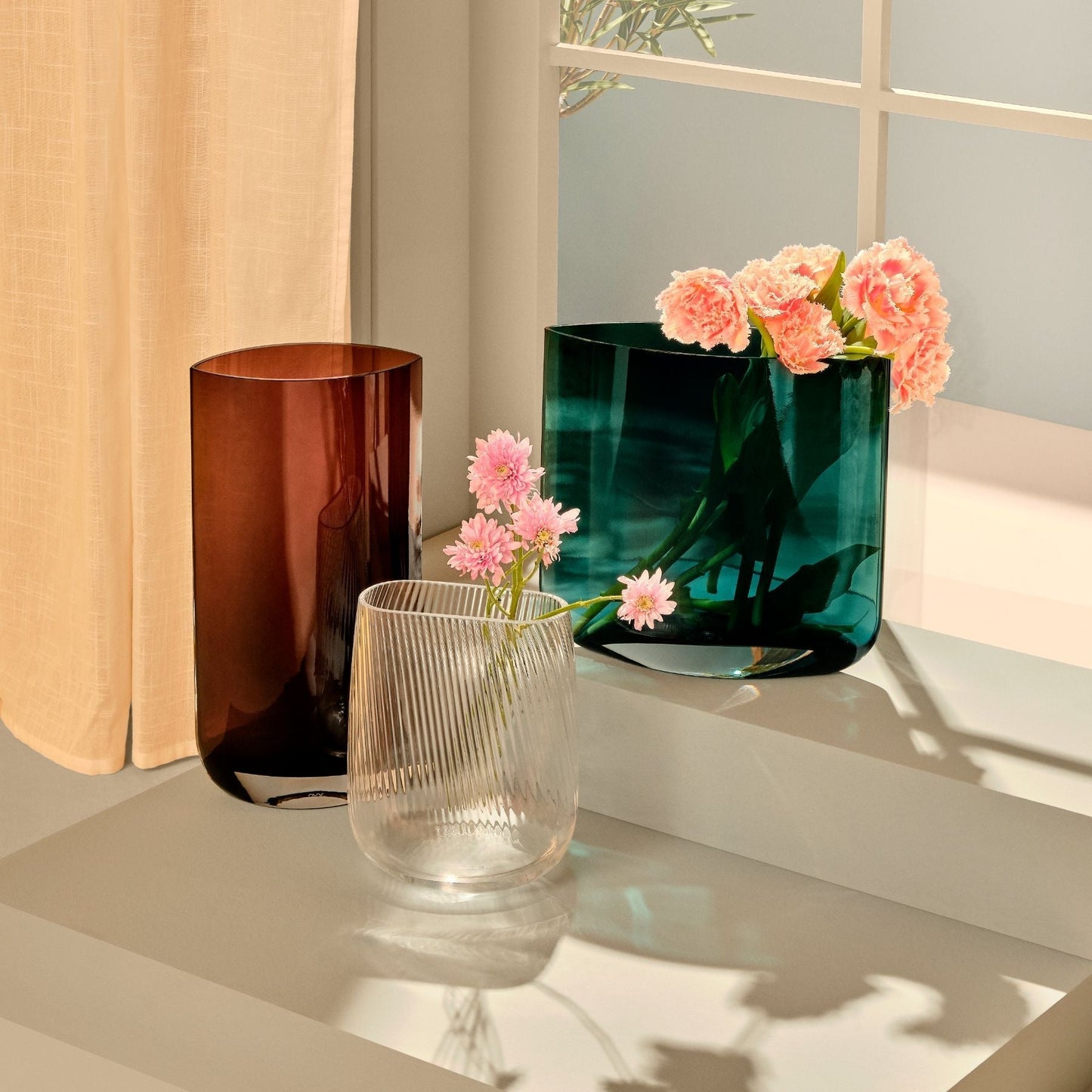 Three vases with flowers on a reflective surface, including a clear vase, a brown vase, and a green vase.