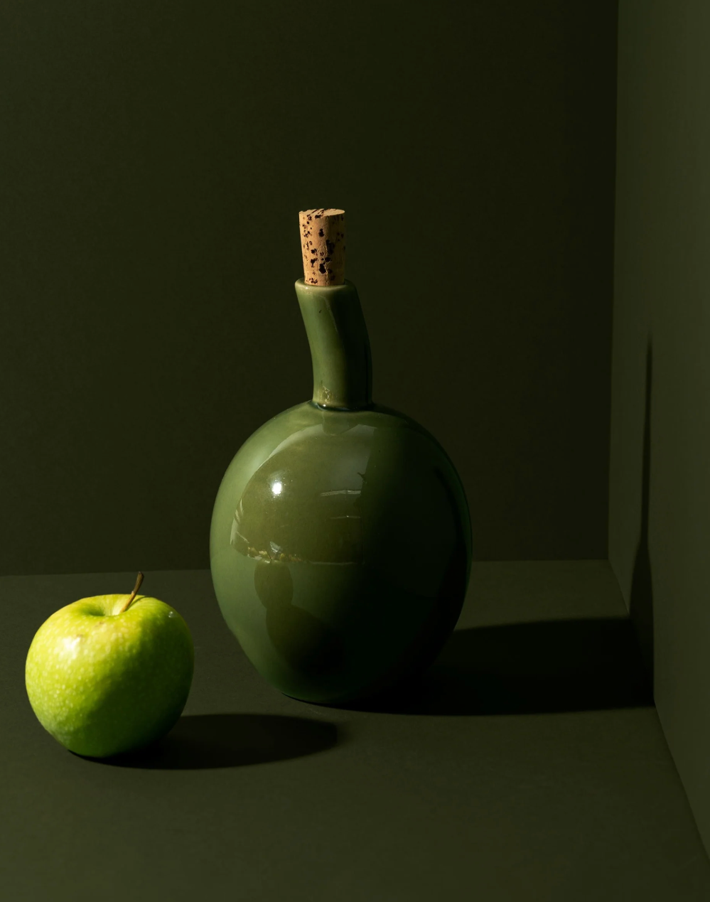 Green ceramic bottle with cork stopper next to a green apple on a dark surface.