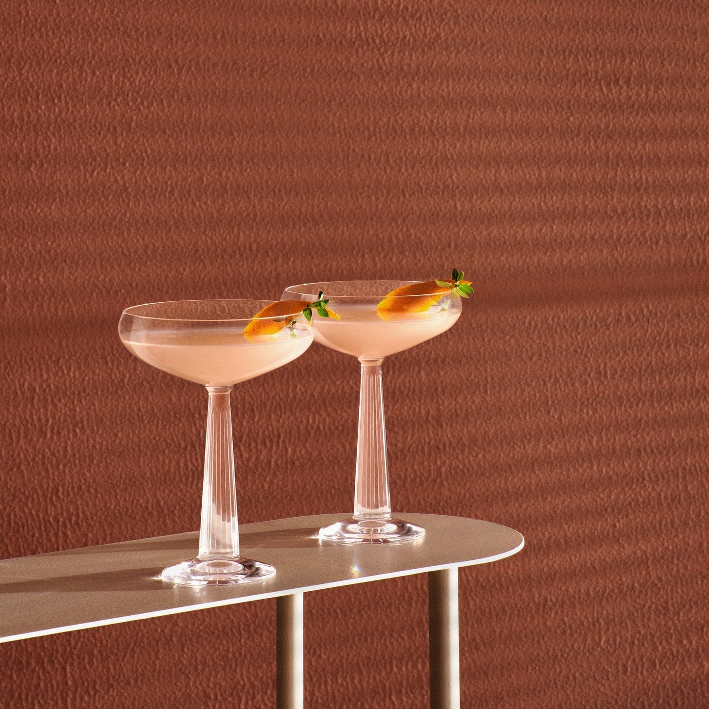 Two cocktail glasses with a pink drink on a metallic surface against a brown textured background