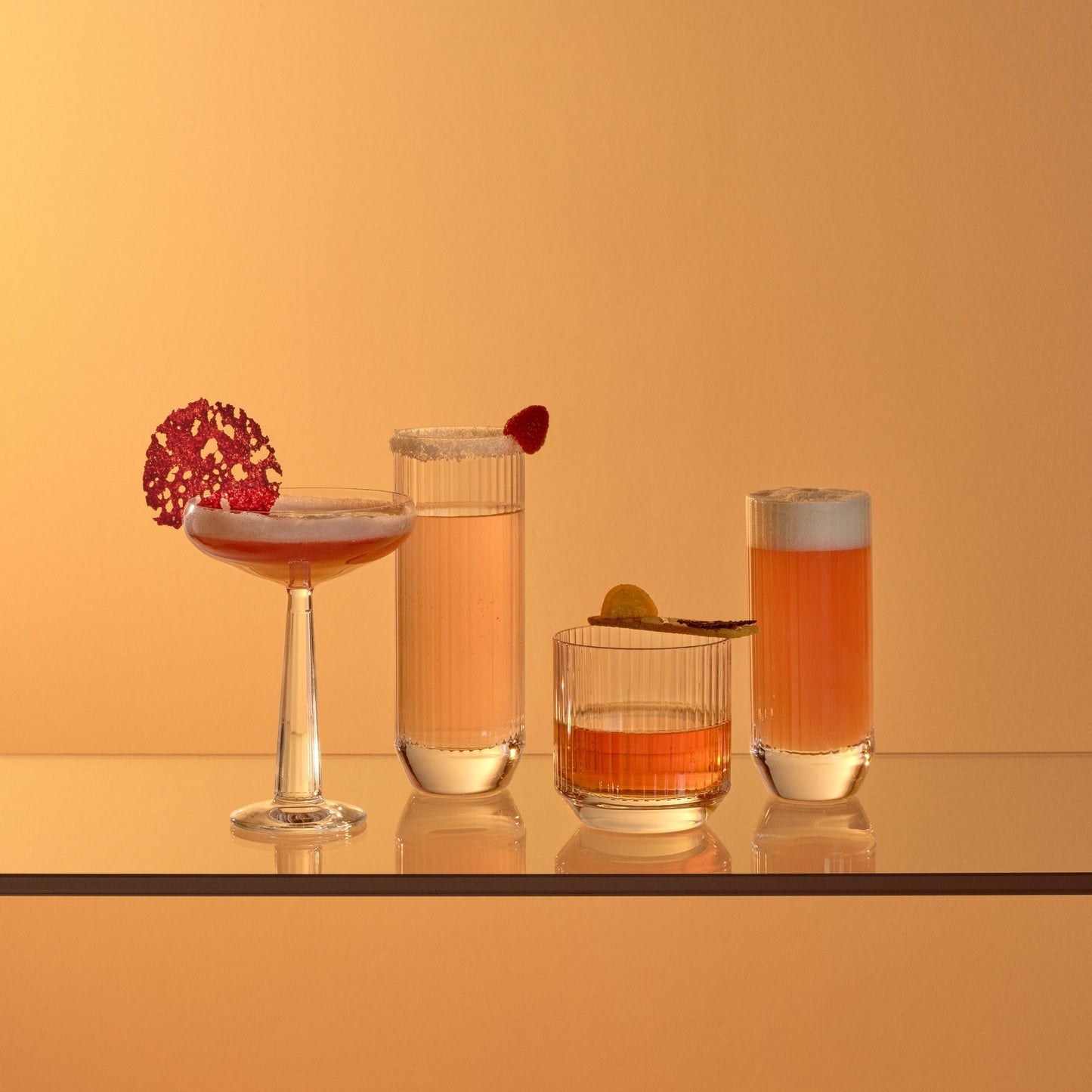 Four cocktails in different glasses on a reflective surface with a warm orange background