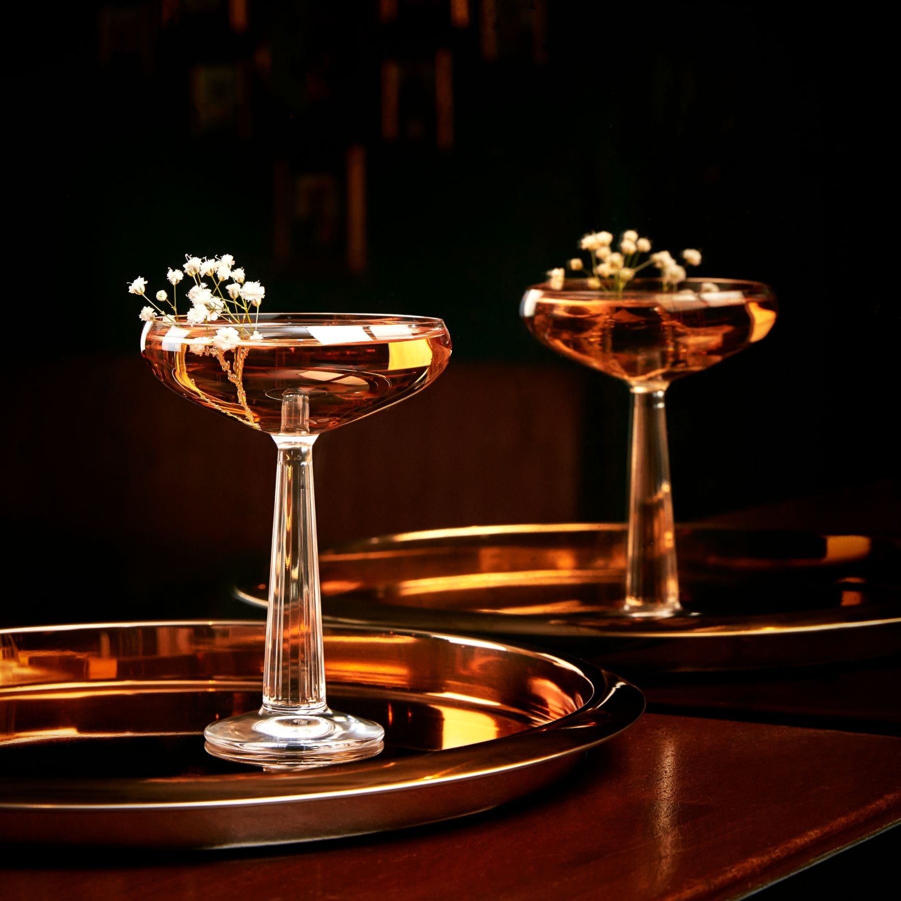 Two cocktail glasses with a golden liquid and small white flowers on a dark background