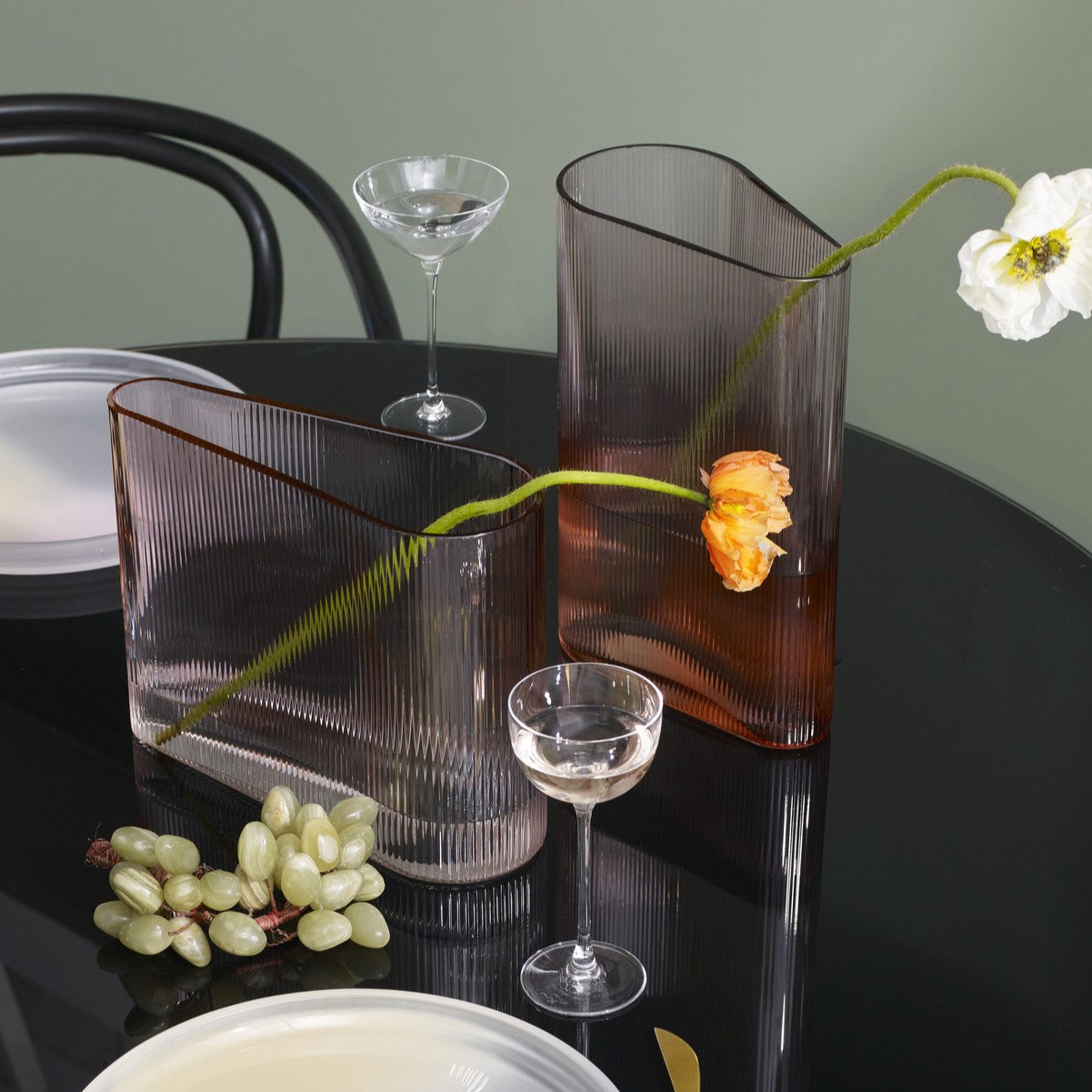 Modern glass vases and glasses on a dark surface with a white flower and grapes.
