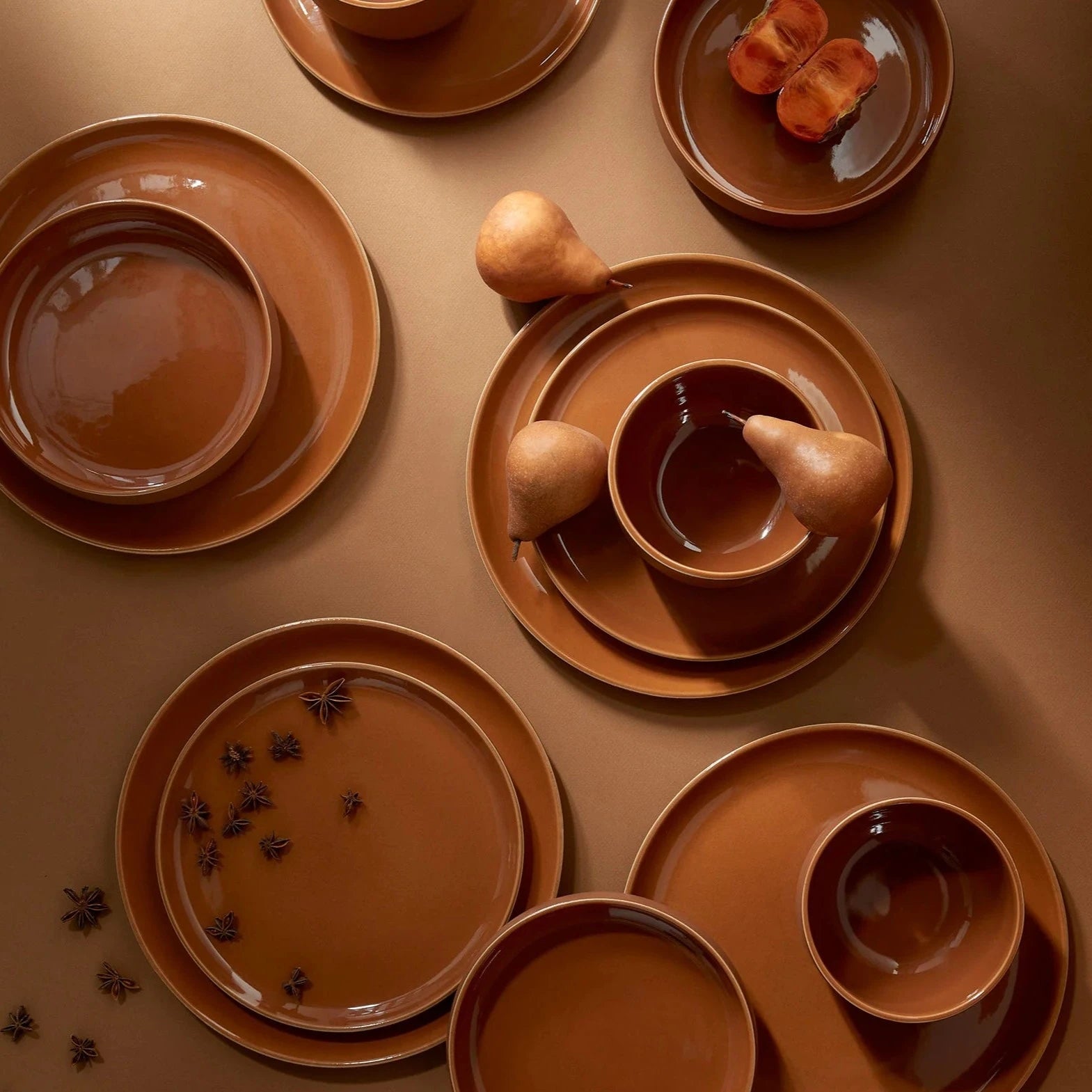 Set of brown ceramic plates and bowls on a matching surface with pears and star anise.