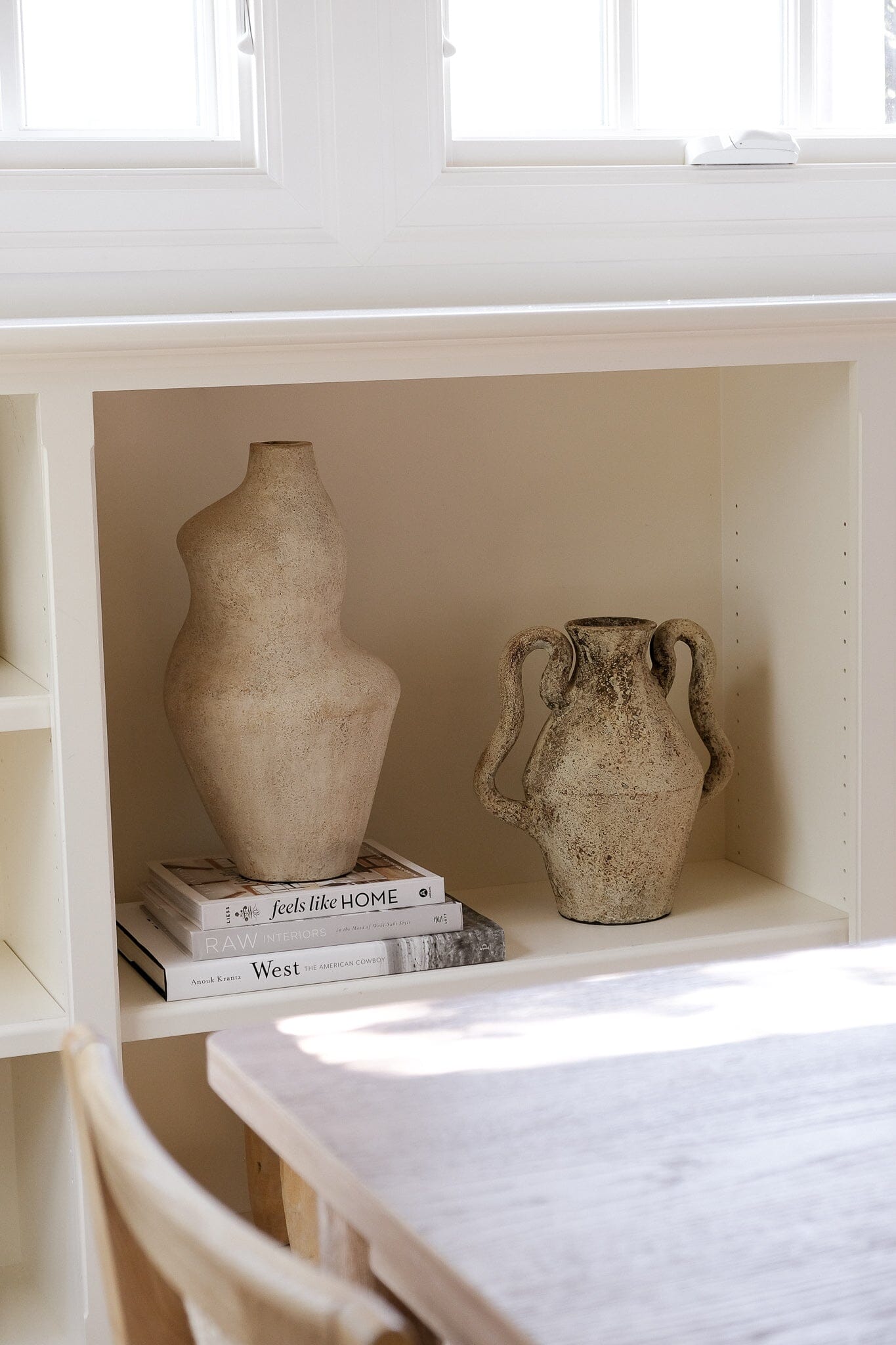Two ceramic vases on a shelf with books underneath, in a bright room.
