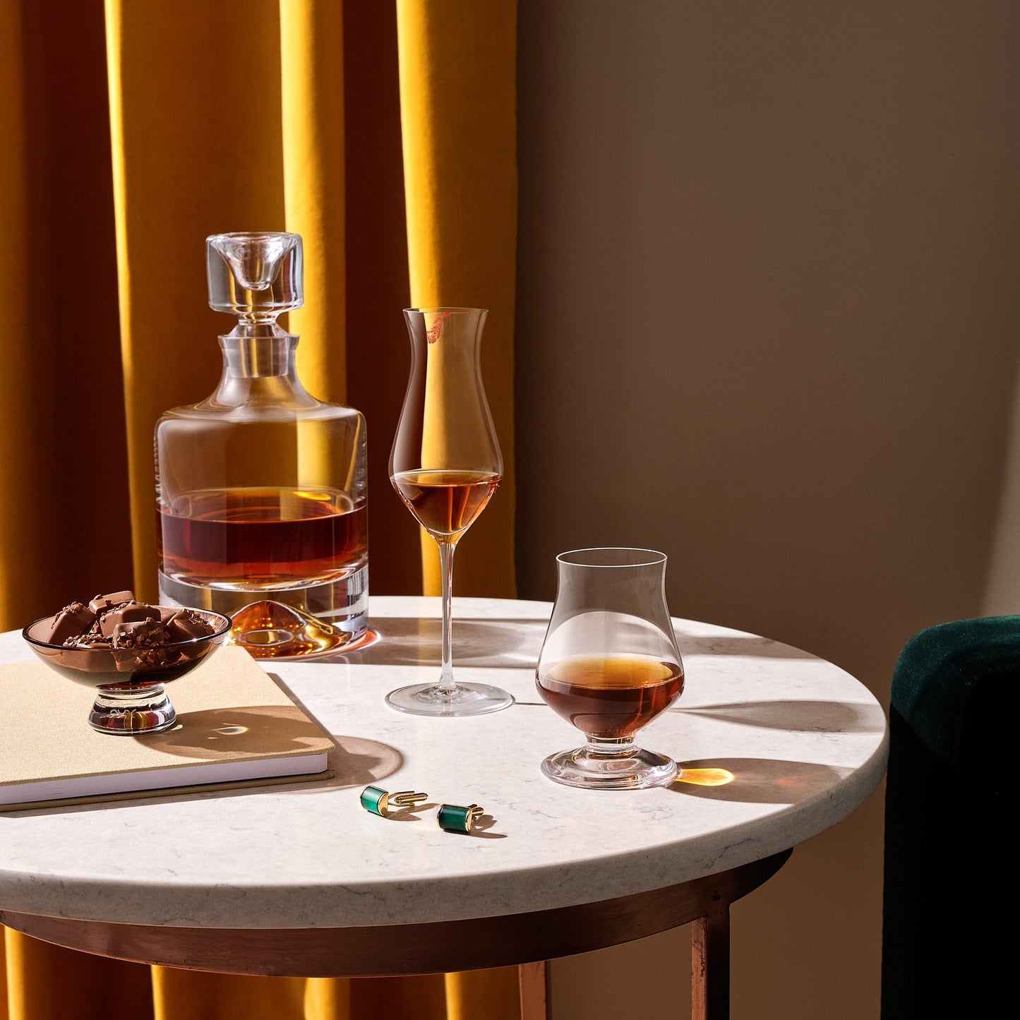 Round table with a decanter, glasses, and snacks against a warm-toned curtain background