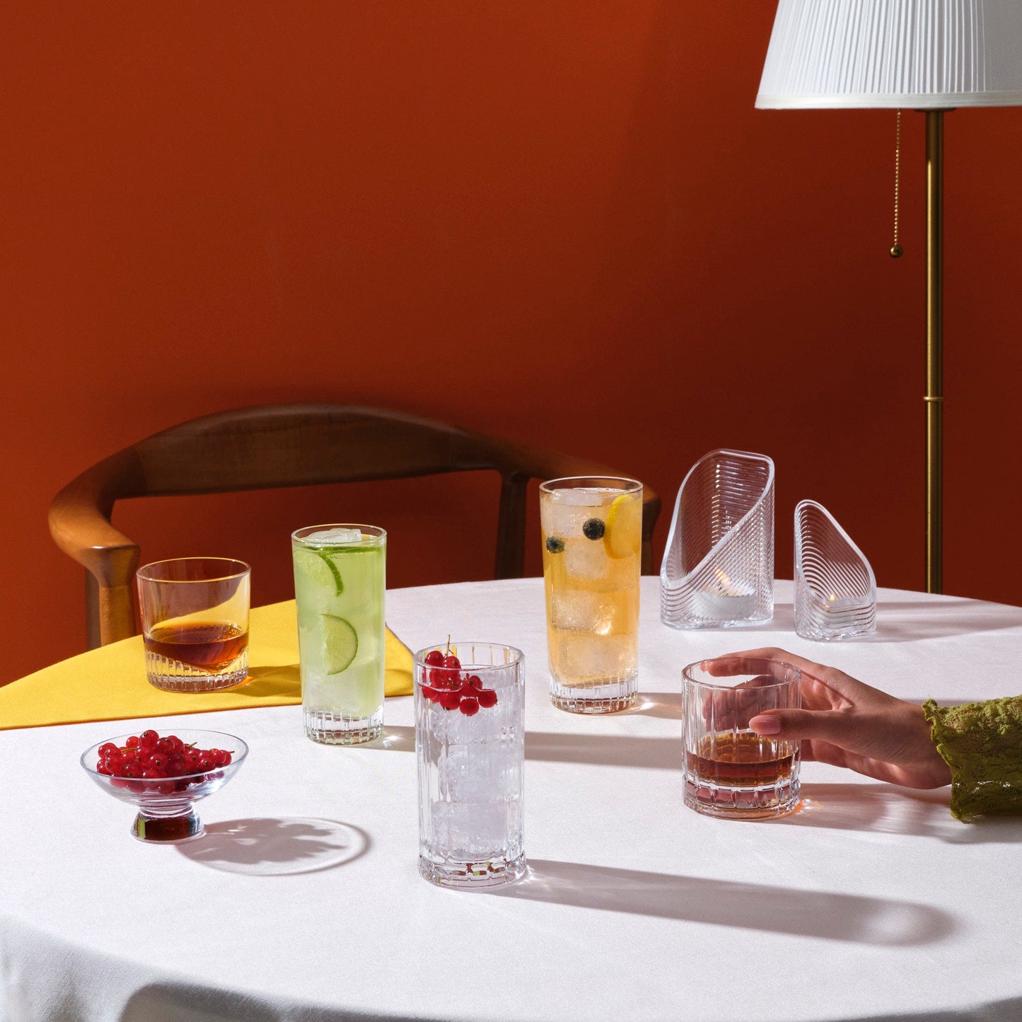Table setting with various glasses of drinks and a hand holding a glass against an orange wall.